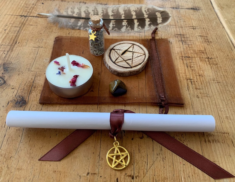 Peut inclure: Une pochette en cuir marron contenant une bougie blanche, une petite bouteille en verre avec un charme d'&eacute;toile dor&eacute;e, un cercle en bois avec un pentagramme grav&eacute;, une plume brune et une pierre brun fonc&eacute;. La pochette est sur une surface en bois.