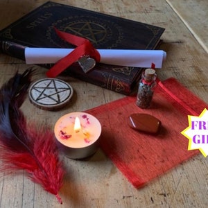 May include: A red and black feather, a small wooden pentagram, a red candle with a flame, a red pouch, a small bottle with a cork, a red stone, a rolled up piece of paper tied with a red ribbon, a book with the word "Spells" on the cover, and a yellow starburst with the words "FREE GIFT" in purple text.
