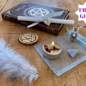 May include: A witchy spell kit with a brown leather-bound book with the word "Spells" on the cover, a white feather, a small wooden circle with a pentagram carved into it, a small glass bottle with a silver star charm, a tealight candle with red wax, a small blue stone, and a blue organza bag.