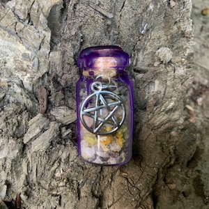 May include: A small clear glass bottle filled with dried yellow flowers and light-colored crystals. The bottle is sealed with a cork and purple wax, and a silver-toned metal pentagram charm is attached to the neck with a piece of twine.