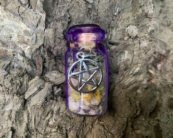 Puede incluir: Una pequeña botella de vidrio transparente llena de flores amarillas secas y cristales claros. La botella está sellada con un corcho y cera morada, y un dije de pentagrama de metal plateado está sujeto al cuello con un cordel.