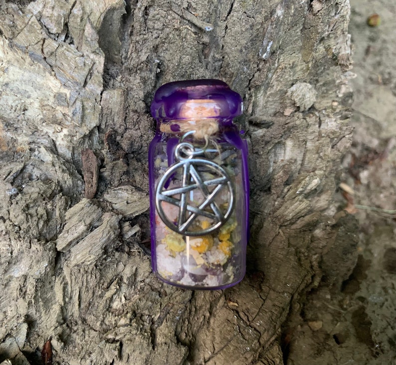 May include: A small clear glass bottle filled with dried yellow flowers and light-coloured crystals. The bottle is sealed with a cork and purple wax, and a silver-toned metal pentagram charm is attached to the neck with a piece of twine.