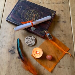 May include: A witchy spell kit with a book titled "Spells", a rolled up scroll, a small bottle with a star charm, a wooden pentagram, a teal and orange feather, a small orange candle, and a carnelian stone in an orange organza bag.