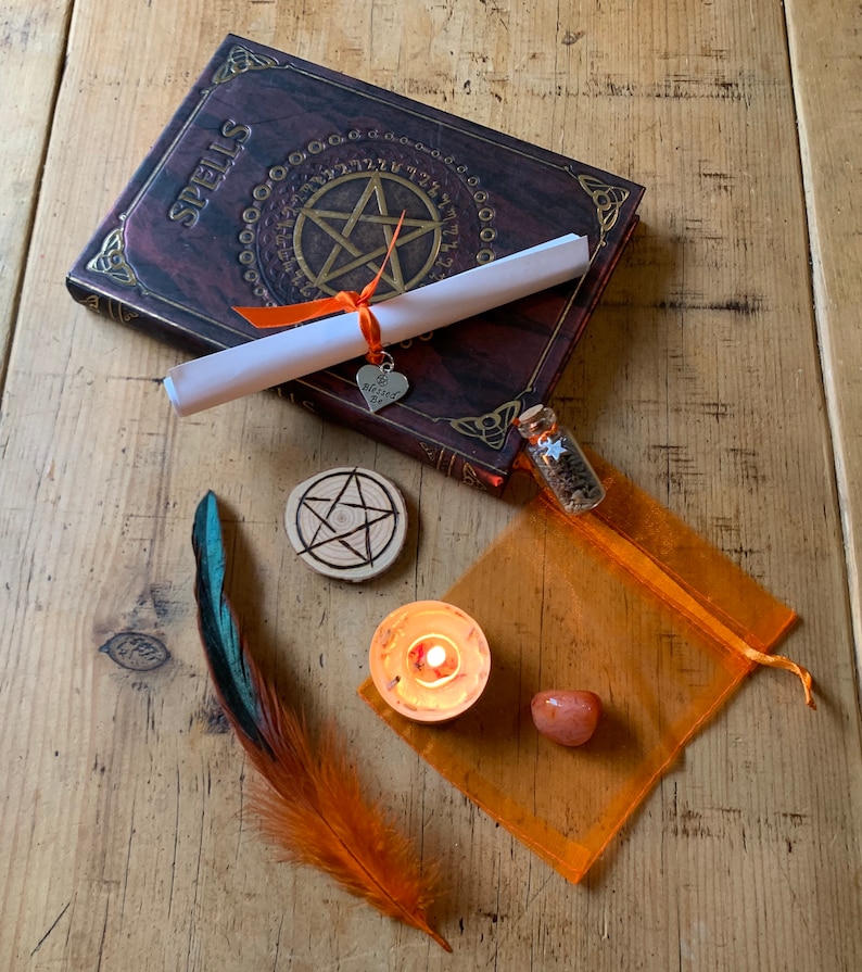 May include: A witchy spell kit with a book titled "Spells", a rolled up scroll, a small bottle with a star charm, a wooden pentagram, a teal and orange feather, a small orange candle, and a carnelian stone in an orange organza bag.