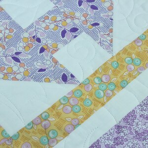 Puede incluir: Una colcha con un dise&ntilde;o de patchwork que presenta una variedad de telas en tonos de morado, amarillo y blanco. La colcha est&aacute; acolchada con un patr&oacute;n de puntadas simple.