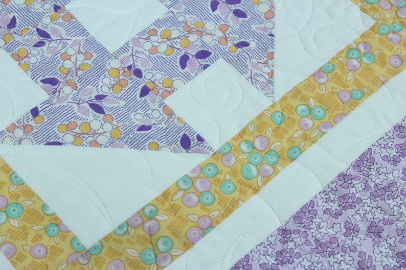 Puede incluir: Una colcha con un dise&ntilde;o de patchwork que presenta una variedad de telas en tonos de morado, amarillo y blanco. La colcha est&aacute; acolchada con un patr&oacute;n de puntadas simple.
