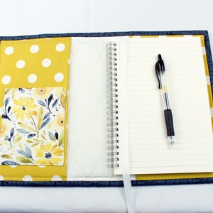 May include: A blue and yellow fabric notebook cover with a floral pocket and a white polka dot design. The cover has a pen holder and a white ribbon bookmark.