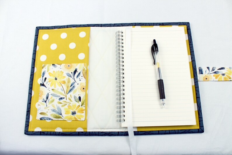 May include: A blue and yellow fabric notebook cover with a floral pocket and a white polka dot design. The cover has a pen holder and a white ribbon bookmark.