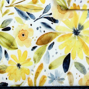 May include: A white fabric with a watercolor floral pattern. The flowers are yellow and orange with blue and green leaves. The fabric has a quilted texture.