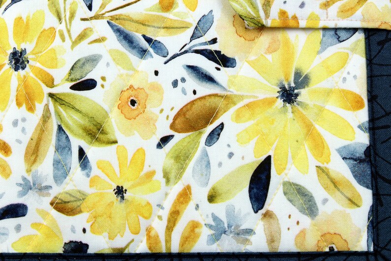 May include: A white fabric with a watercolor floral pattern. The flowers are yellow and orange with blue and green leaves. The fabric has a quilted texture.