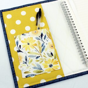 May include: A yellow and white polka dot notebook cover with a floral fabric pocket and a blue pen. The notebook has a spiral binding.