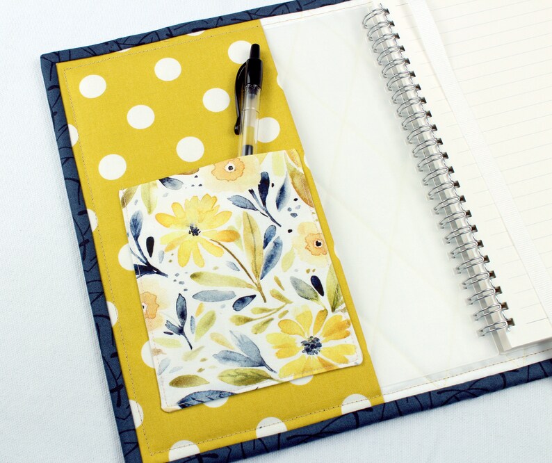 May include: A yellow and white polka dot notebook cover with a floral fabric pocket and a blue pen. The notebook has a spiral binding.