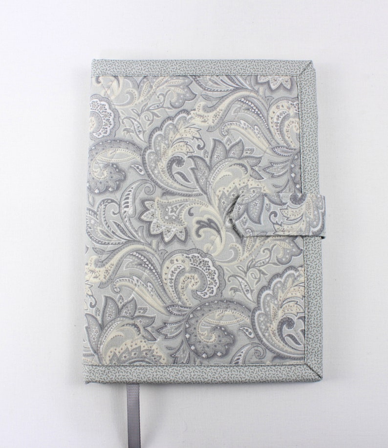 Pu&ograve; includere: Una copertina per libro in tessuto con motivo paisley grigio e bianco con un cinturino in tessuto grigio.