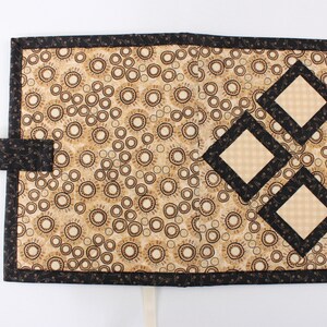 Puede incluir: Una cubierta de libro de tela negra y beige con un patr&oacute;n decorativo de c&iacute;rculos y un dise&ntilde;o de tres cuadrados superpuestos con un patr&oacute;n de cuadros. La cubierta tiene una correa de tela negra y una cinta blanca.