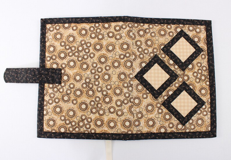 Puede incluir: Una cubierta de libro de tela negra y beige con un patr&oacute;n decorativo de c&iacute;rculos y un dise&ntilde;o de tres cuadrados superpuestos con un patr&oacute;n de cuadros. La cubierta tiene una correa de tela negra y una cinta blanca.