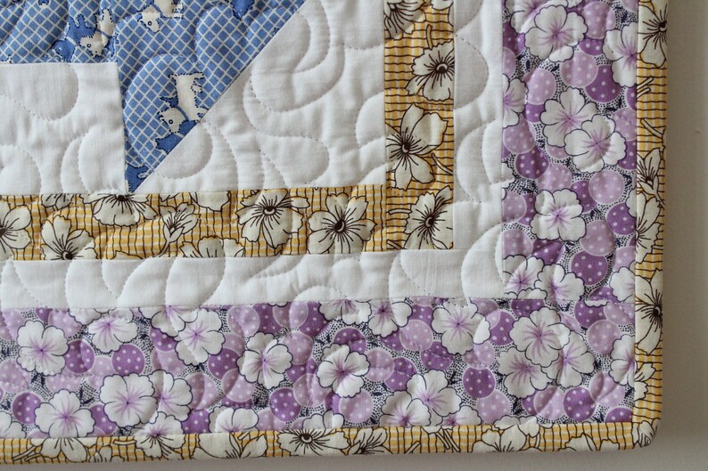 May include: Close-up of a quilt corner with a white background and a border of yellow and purple floral fabric. The quilt is quilted with a wavy pattern.