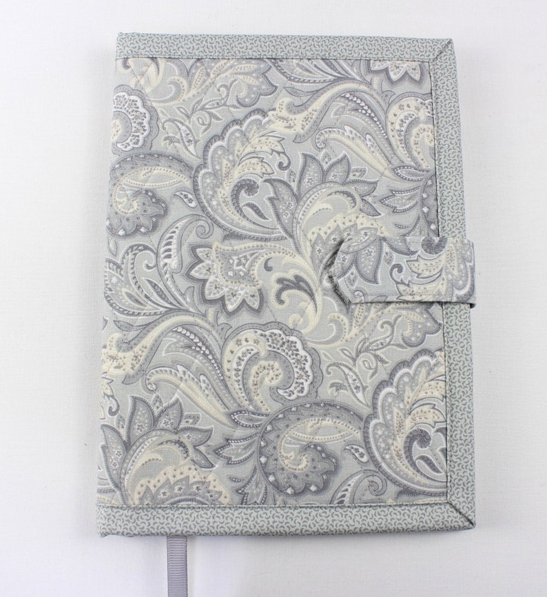 Pu&ograve; includere: Una copertina per libro in tessuto con motivo paisley grigio e bianco con chiusura a lembo in tessuto grigio e un segnalibro in nastro grigio.
