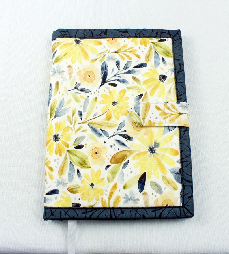 May include: A blue and yellow floral fabric book cover with a strap closure. The cover has a watercolor-style design with yellow flowers and blue leaves.
