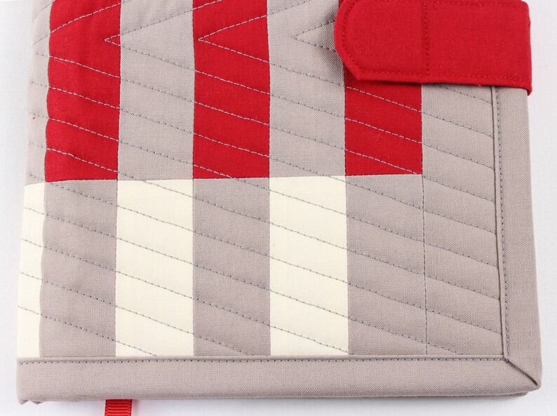 May include: A grey and red quilted fabric notebook cover with a red fabric closure strap. The cover has a chequered pattern with diagonal stitching.