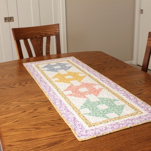 May include: A colourful quilted table runner with a geometric pattern in shades of yellow, pink, green, and blue. The runner is placed on a wooden table with two chairs.