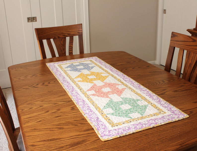 May include: A colourful quilted table runner with a geometric pattern in shades of yellow, pink, green, and blue. The runner is placed on a wooden table with two chairs.