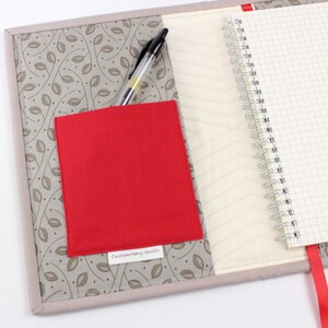 May include: A grey fabric notebook cover with a red fabric pocket and a black pen. The cover has a leaf pattern and the pocket has a label that says "Custom Mary Quilts".