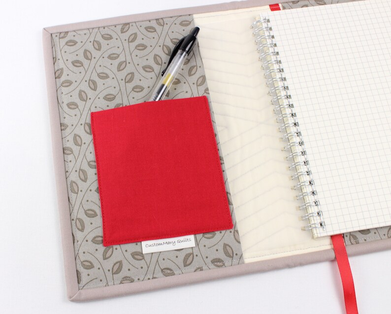 May include: A grey fabric notebook cover with a red fabric pocket and a black pen. The cover has a leaf pattern and the pocket has a label that says "Custom Mary Quilts".