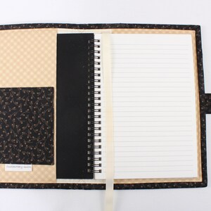 Puede incluir: Un cuaderno de espiral cubierto de tela negra y beige. El cuaderno tiene un bolsillo en la portada y un marcador de cinta. El texto "Custom Made Goods" est&aacute; impreso en el bolsillo.