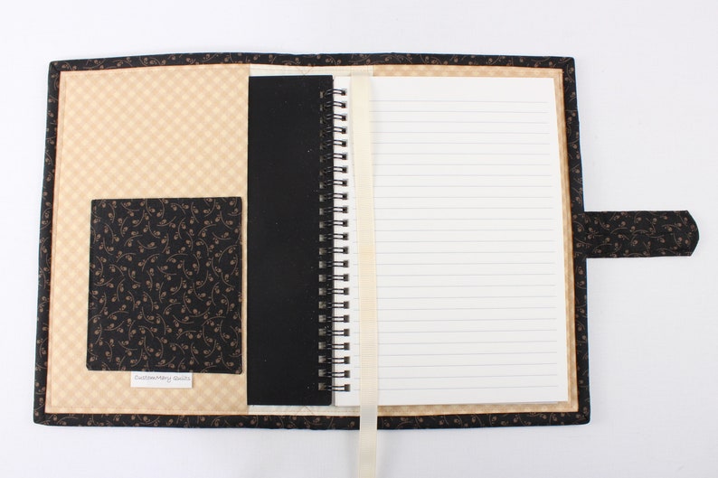 Puede incluir: Un cuaderno de espiral cubierto de tela negra y beige. El cuaderno tiene un bolsillo en la portada y un marcador de cinta. El texto "Custom Made Goods" est&aacute; impreso en el bolsillo.