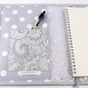 Pu&ograve; includere: Una copertina per quaderno in tessuto grigio e bianco a pois con una tasca in tessuto paisley e una penna. La copertina ha un'etichetta bianca con il testo "Customary Quilts".