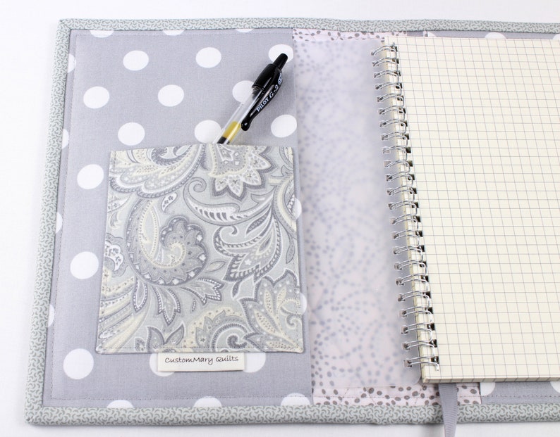 Pu&ograve; includere: Una copertina per quaderno in tessuto grigio e bianco a pois con una tasca in tessuto paisley e una penna. La copertina ha un'etichetta bianca con il testo "Customary Quilts".