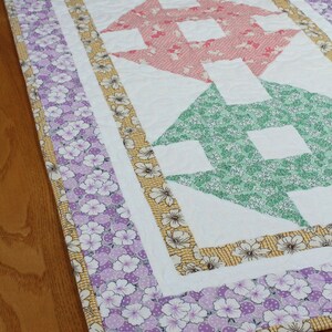 May include: A white table runner with a patchwork design featuring a series of colourful arrows. The arrows are made from different floral patterned fabrics in shades of blue, yellow, pink, green, and purple. The runner has a border of purple floral fabric.