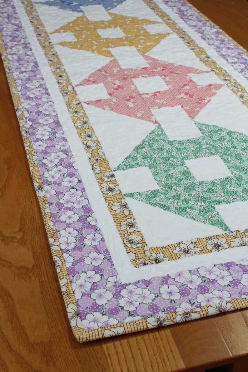 May include: A white table runner with a patchwork design featuring a series of colourful arrows. The arrows are made from different floral patterned fabrics in shades of blue, yellow, pink, green, and purple. The runner has a border of purple floral fabric.