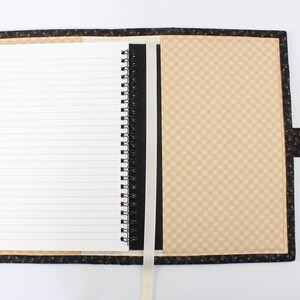 Puede incluir: Un cuaderno negro y marr&oacute;n con un patr&oacute;n a cuadros en la portada. El cuaderno tiene una encuadernaci&oacute;n en espiral negra y un marcador de cinta blanco. El cuaderno est&aacute; abierto en una p&aacute;gina en blanco con papel rayado.