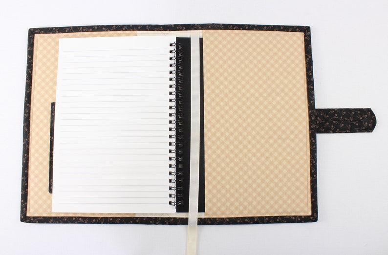 Puede incluir: Un cuaderno negro y marr&oacute;n con un patr&oacute;n a cuadros en la portada. El cuaderno tiene una encuadernaci&oacute;n en espiral negra y un marcador de cinta blanco. El cuaderno est&aacute; abierto en una p&aacute;gina en blanco con papel rayado.