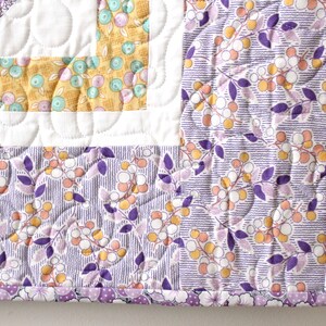 Puede incluir: Una colcha con estampado floral de color morado y amarillo sobre fondo blanco. La colcha est&aacute; acolchada con un patr&oacute;n de espiral. La colcha est&aacute; ribeteada con un ribete morado y blanco.
