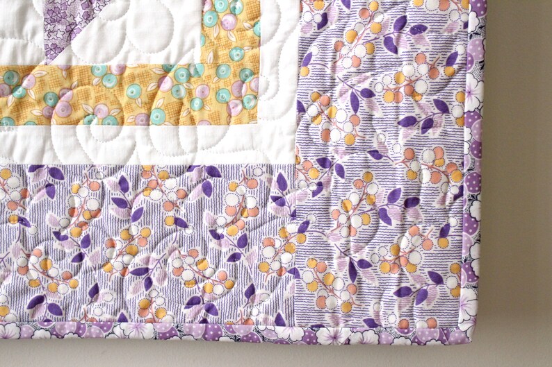 Puede incluir: Una colcha con estampado floral de color morado y amarillo sobre fondo blanco. La colcha est&aacute; acolchada con un patr&oacute;n de espiral. La colcha est&aacute; ribeteada con un ribete morado y blanco.