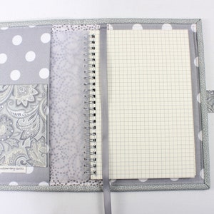 Pu&ograve; includere: Una copertina per quaderno a pois grigi e bianchi con una tasca paisley e un quaderno a spirale argentato. La copertina ha un'etichetta che recita "Custom Made by Suzi".