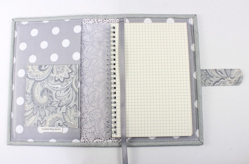 Pu&ograve; includere: Una copertina per quaderno a pois grigi e bianchi con una tasca paisley e un quaderno a spirale argentato. La copertina ha un'etichetta che recita "Custom Made by Suzi".