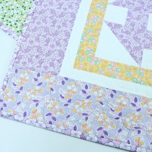 Puede incluir: Una colcha floral de color morado y amarillo con un cuadrado central blanco y un patr&oacute;n geom&eacute;trico. La colcha est&aacute; hecha con diferentes telas y tiene una textura acolchada.