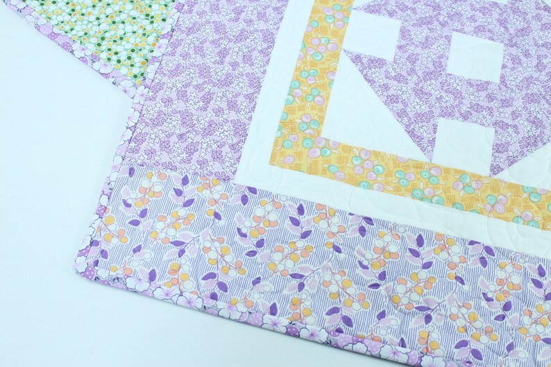 Puede incluir: Una colcha floral de color morado y amarillo con un cuadrado central blanco y un patr&oacute;n geom&eacute;trico. La colcha est&aacute; hecha con diferentes telas y tiene una textura acolchada.