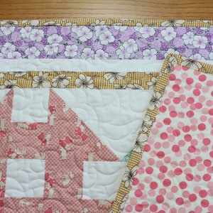 May include: A pink and white quilt with a house-shaped design in the centre. The quilt is made with different fabrics, including a floral print, a polka dot print, and a plain white fabric. The quilt is quilted with a wavy pattern.