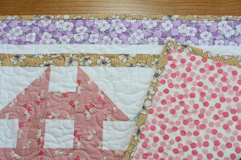 May include: A pink and white quilt with a house-shaped design in the centre. The quilt is made with different fabrics, including a floral print, a polka dot print, and a plain white fabric. The quilt is quilted with a wavy pattern.
