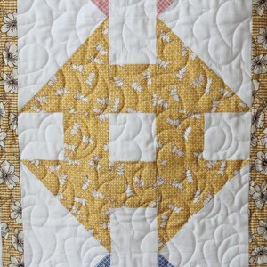 May include: A quilt block featuring a yellow and white floral pattern in the centre, surrounded by a white background with curved quilting lines. The block is bordered by strips of fabric with purple and white floral patterns.