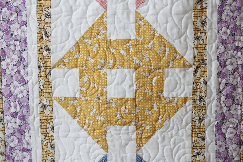 May include: A quilt block featuring a yellow and white floral pattern in the centre, surrounded by a white background with curved quilting lines. The block is bordered by strips of fabric with purple and white floral patterns.