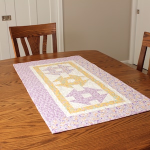 Puede incluir: Un camino de mesa acolchado morado y amarillo con un patr&oacute;n geom&eacute;trico. El camino de mesa est&aacute; sobre una mesa de madera.