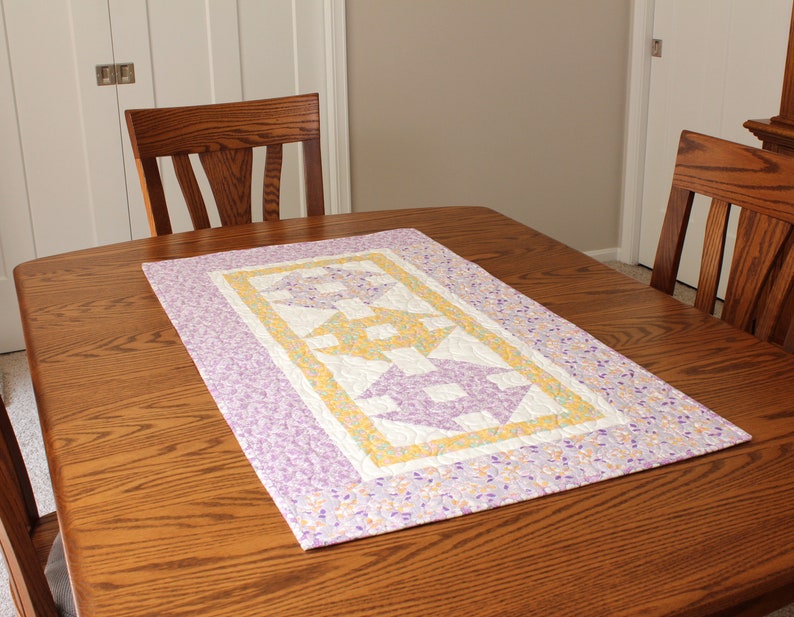 Puede incluir: Un camino de mesa acolchado morado y amarillo con un patr&oacute;n geom&eacute;trico. El camino de mesa est&aacute; sobre una mesa de madera.
