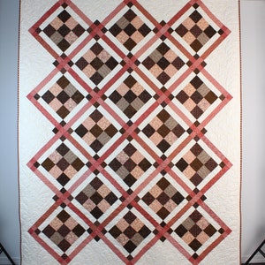 May include: A quilt with a diamond pattern in shades of brown, pink, and white. The quilt is made with a variety of fabrics and has a white background.