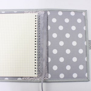 Pu&ograve; includere: Una copertina per quaderno a pois grigi e bianchi con una tasca trasparente in plastica per una penna. La copertina ha una patta in tessuto grigio con un motivo paisley.