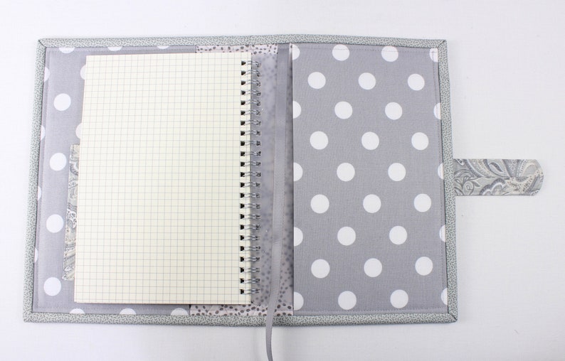 Pu&ograve; includere: Una copertina per quaderno a pois grigi e bianchi con una tasca trasparente in plastica per una penna. La copertina ha una patta in tessuto grigio con un motivo paisley.